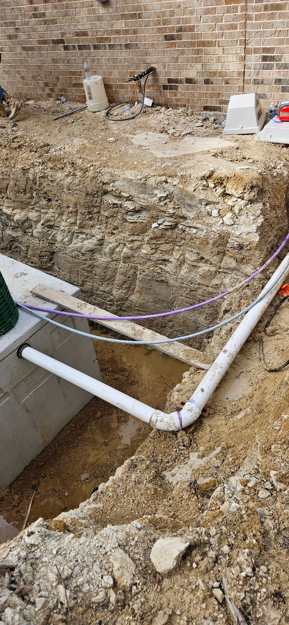 Excavation pit with white PVC pipes and blue tubing against a brick foundation wall, showing soil layers and construction debris