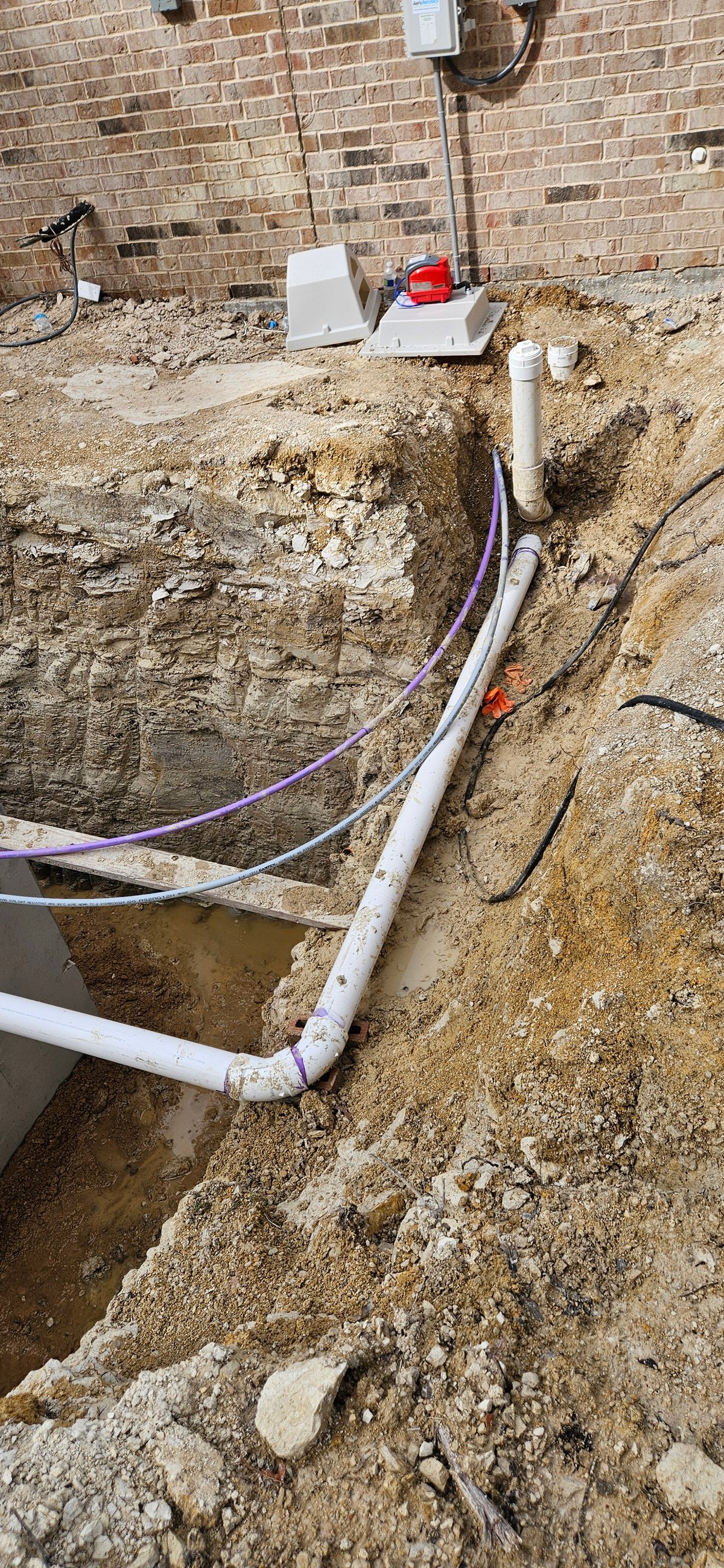 Excavated trench with white PVC pipes and purple electrical conduit along brick foundation wall